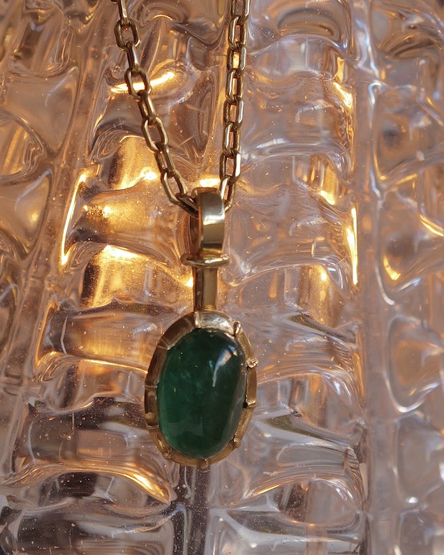 Oval emerald cabochon pendant in a hand-formed gold bezel, hanging from a gold chain in front of textured glass